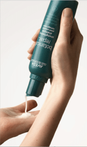 A person dispenses a white liquid from a dark green Aveda bottle into their hand.